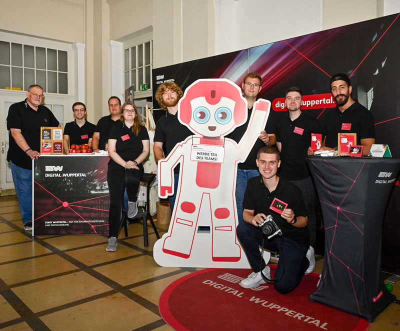 Azubis und Ausbilder aus dem Team 402 stehen vor dem Messestand, in der Mitte steht ein Pappaufsteller des Roboter-Maskottchens Yanny.